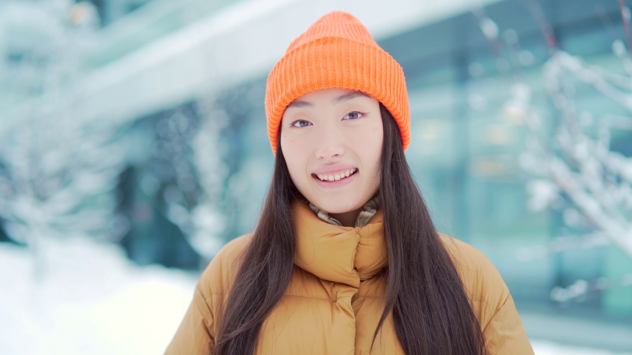 画像年轻的亚洲妇女站在雪地背景在现代城市街道建筑在寒冷的天气看着相机。面对快乐微笑的美丽女孩在冬天的户外视频素材