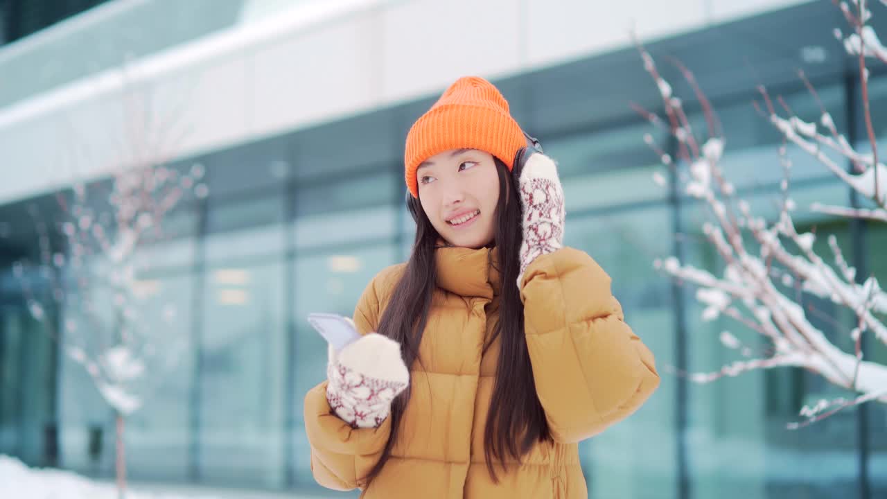 年轻美丽的亚洲女子在冬天的雪天里戴着耳机听音乐，用手机上的在线应用程序，视频素材