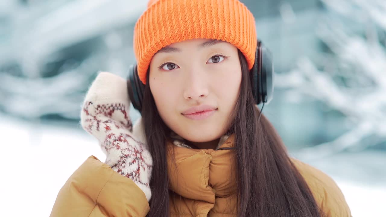 年轻美丽的亚洲女子在冬天的雪天里戴着耳机听音乐，用手机上的在线应用程序，视频素材