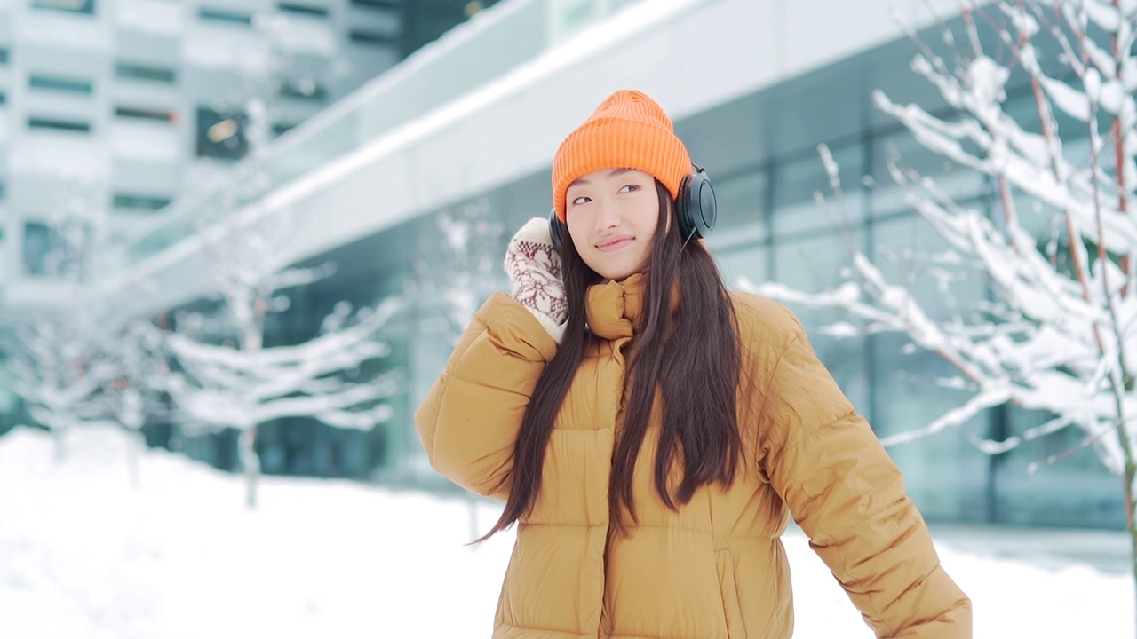 年轻美丽的亚洲女子在冬天的雪天里戴着耳机听音乐，用手机上的在线应用程序，视频素材