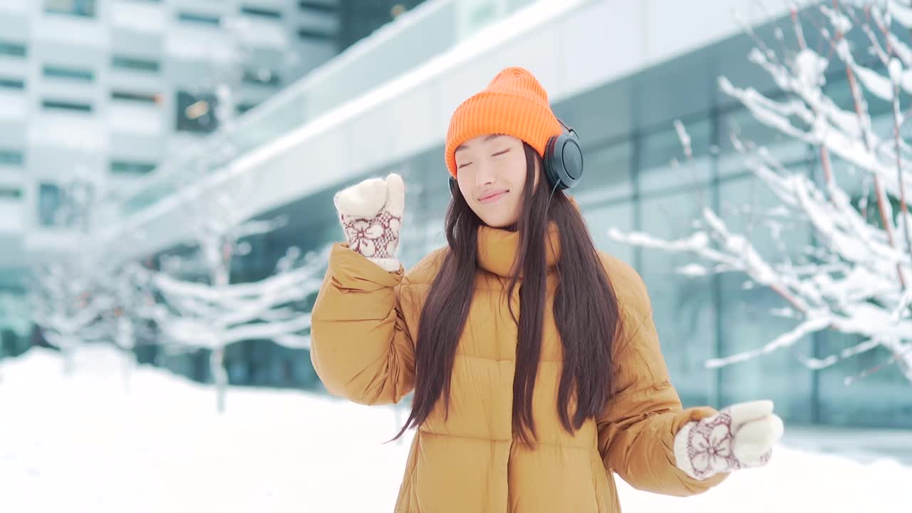 年轻美丽的亚洲女子在冬天的雪天里戴着耳机听音乐，用手机上的在线应用程序，视频素材