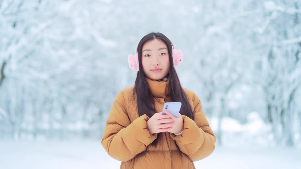 快乐快乐有趣的亚洲少女学生在一个下雪的冬天在公园里散步听音乐。有手机的年轻女子喜欢在寒冷的天气里跳舞。视频素材