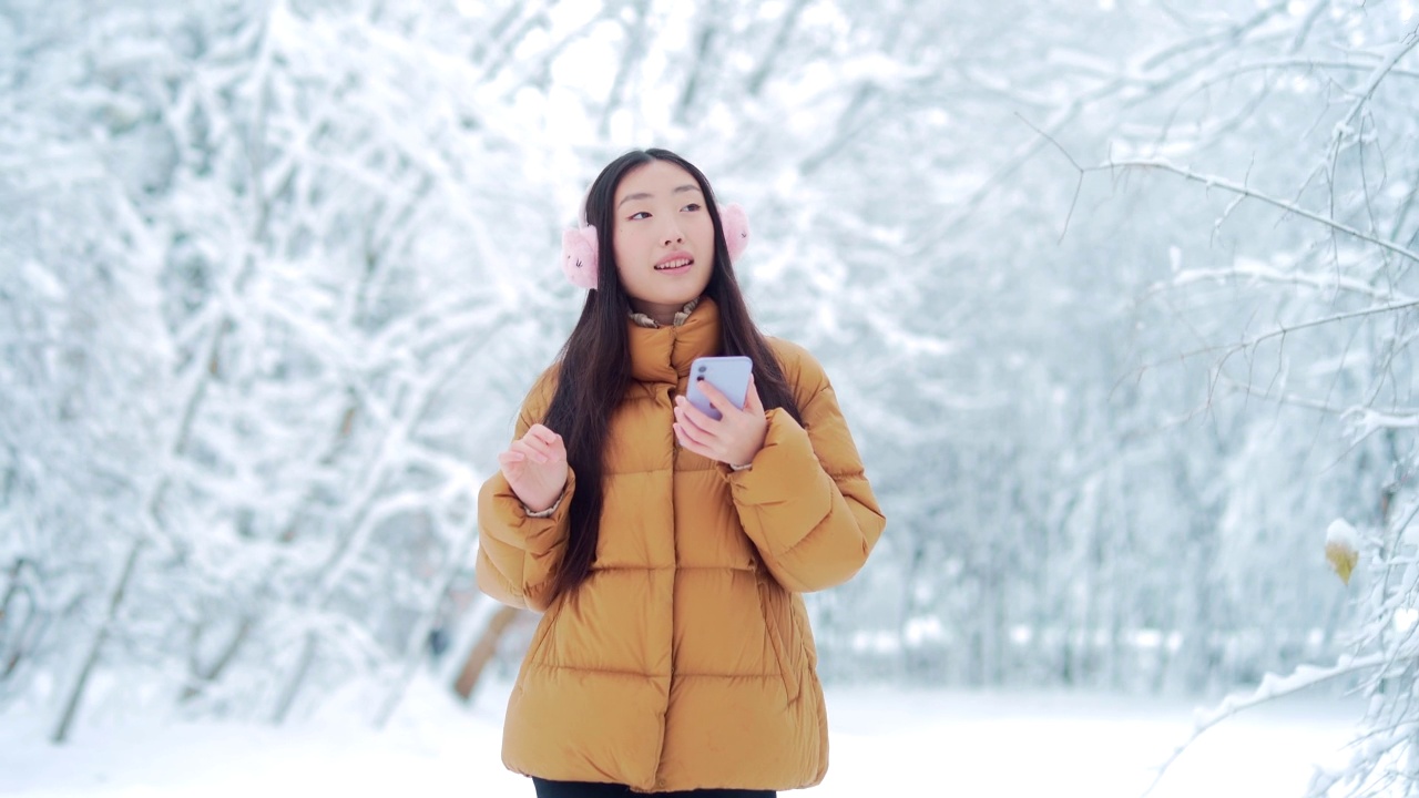 快乐快乐有趣的亚洲少女学生在一个下雪的冬天在公园里散步听音乐。有手机的年轻女子喜欢在寒冷的天气里跳舞。视频素材