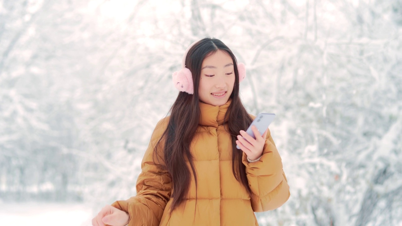 快乐快乐有趣的亚洲少女学生在一个下雪的冬天在公园里散步听音乐。有手机的年轻女子喜欢在寒冷的天气里跳舞。视频素材