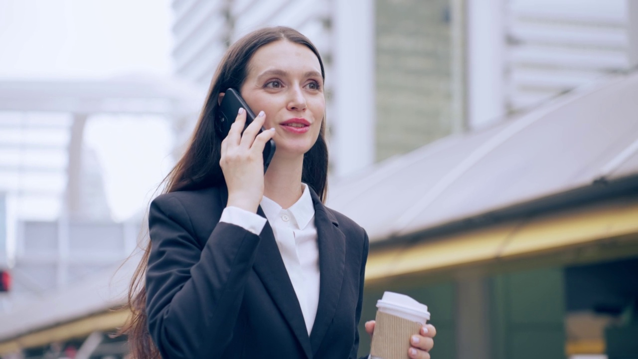 白人年轻女商人在城市里散步时用智能手机说话。迷人美丽的女员工在户外与同事端着咖啡，用手机自信地交流。视频素材