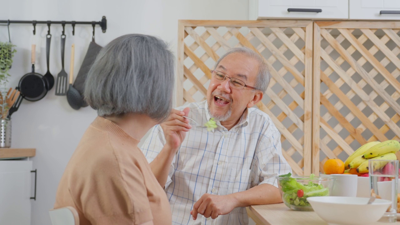 亚洲老年夫妇在家里的厨房里一起吃沙拉。幸福的家庭，有魅力的年长成熟的夫妻坐在餐桌上享受退休生活，午餐吃健康的食物在家里。视频素材