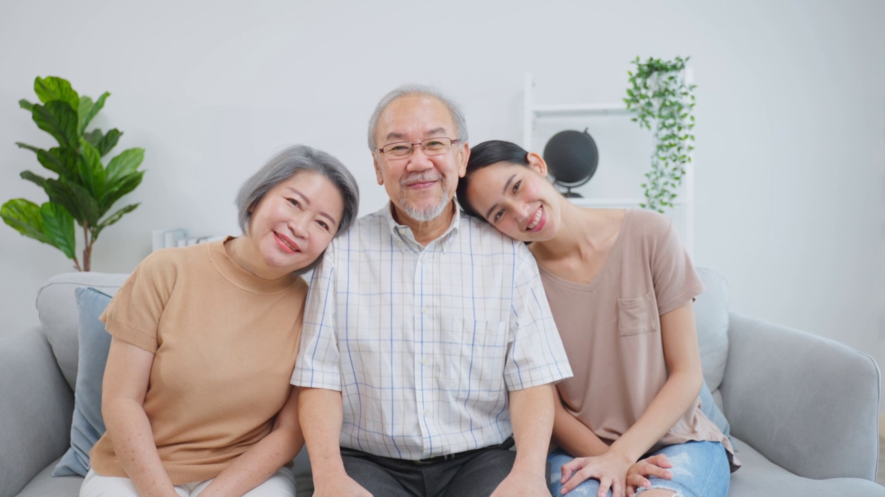 画像的亚洲高级老夫妇坐在年轻的女儿在房子。迷人美丽的孙女拜访年迈成熟的爷爷奶奶然后微笑着，在家里的客厅一起看镜头视频素材