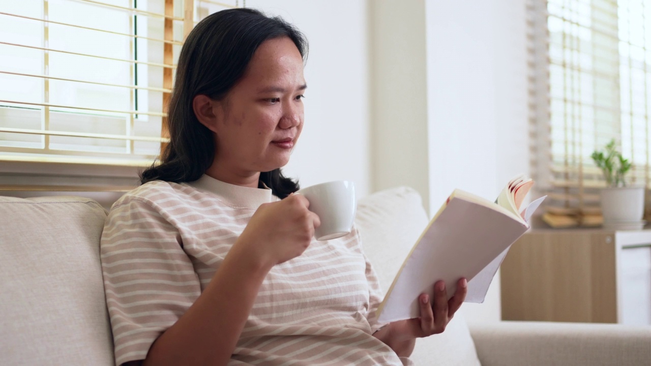 快乐的女人周末坐在沙发上看书，喝咖啡或茶在家里的客厅视频素材