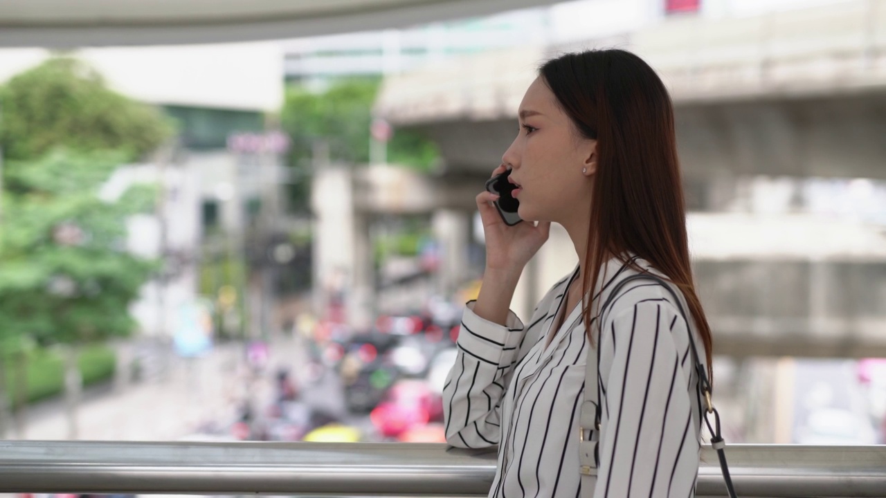 聪明、休闲、酷的亚洲年轻迷人的女商人正在用智能手机给她的朋友打电话，告诉她曼谷市一个商业区的糟糕交通状况，她可能会迟到视频素材