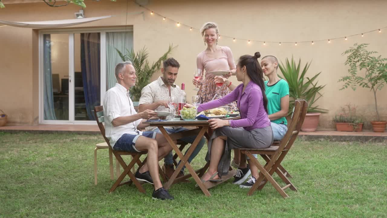 夏天，朋友们在自家花园共进晚餐，分享美食——金发女人倒玫瑰酒庆祝友谊视频素材