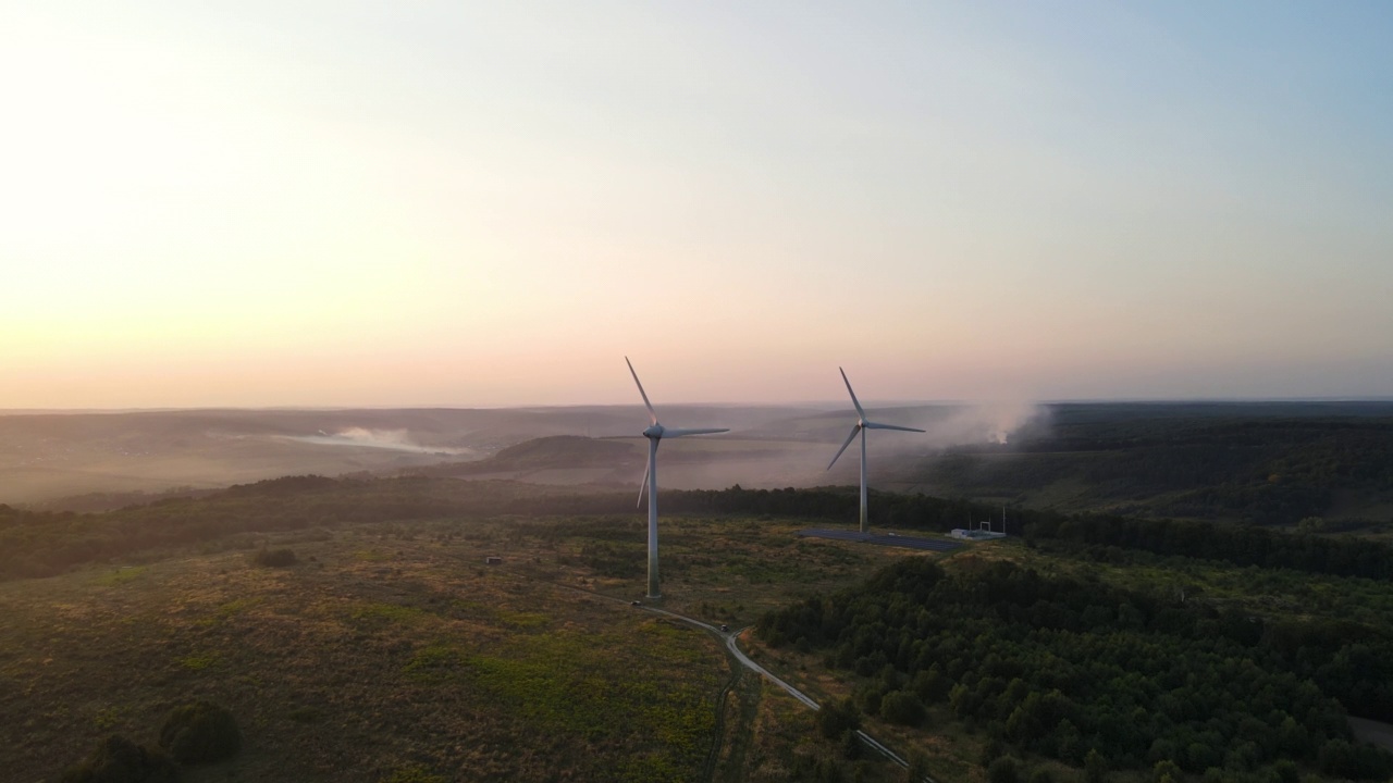 两个旋转叶片的风车的空中全景，工作的风力涡轮机在美丽的日落林地，无人机向前飞行视频素材