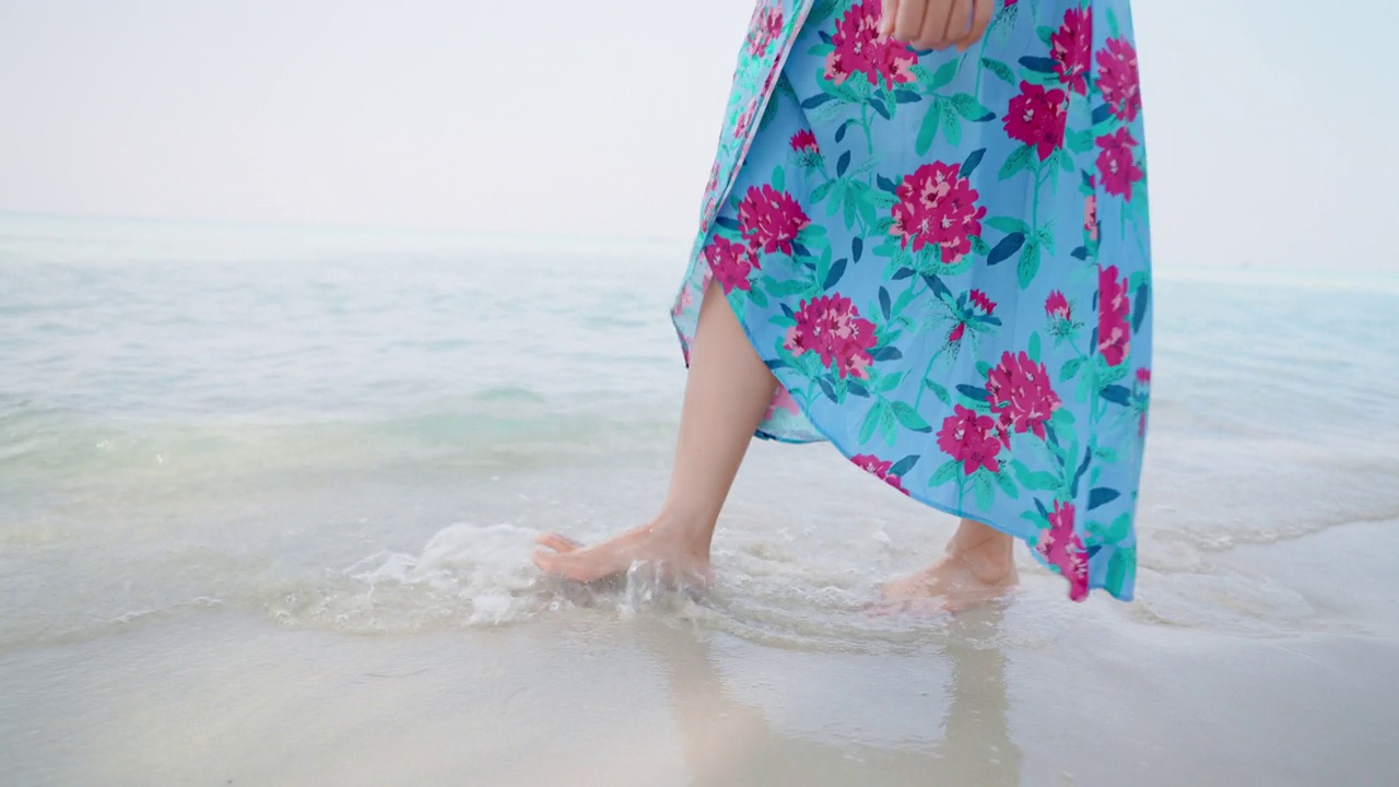 女性光着脚穿着蓝色的裙子走在岛上的海滩上，清澈的海水波浪，溅起的水打在她的腿上，独自旅行，夏季旅行，热带气候，侧视图视频素材