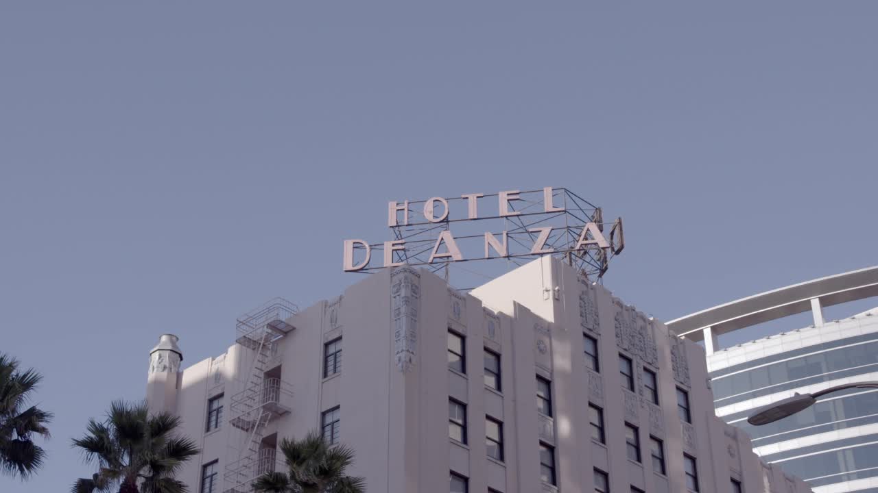 从写着“hotel de anza”的酒店标牌拉回并向下平移到广角的高层建筑，酒店，办公楼和下面的城市街道。墙上可见壁画。汽车在街上行驶。棕榈树可见视频素材