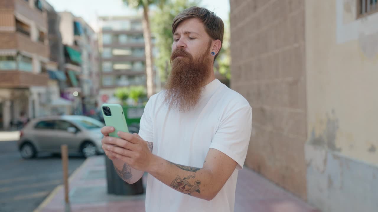 年轻的红发男子微笑着自信地在街上用智能手机自拍视频素材