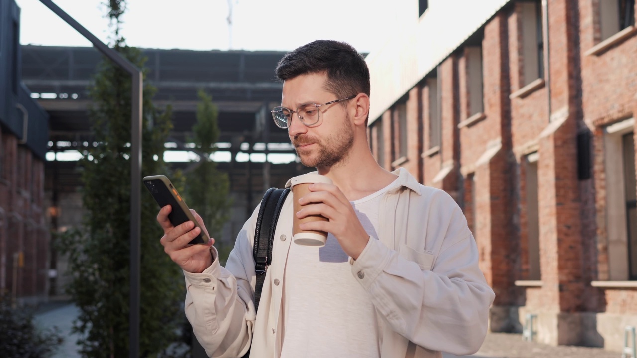 特写年轻自信的英俊男子走在户外，喝着咖啡和阅读短信的手机。迷人的男性站在城外的街道上用手机聊天，一边打短信。视频素材