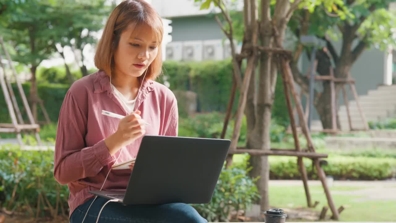 年轻的亚洲女孩戴着耳机，用笔记本电脑上网学习，坐在公园里写笔记本。视频素材