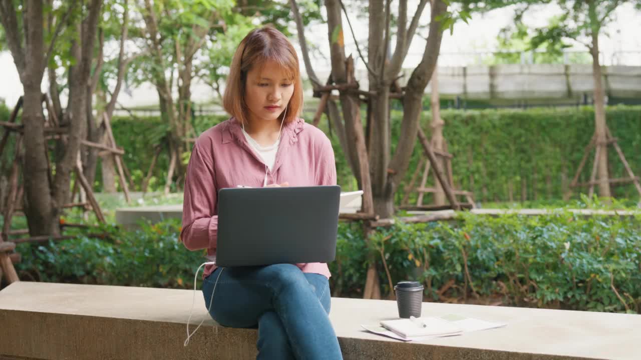 年轻的亚洲女孩戴着耳机，用笔记本电脑上网学习，坐在公园里在平板电脑上写字。视频素材