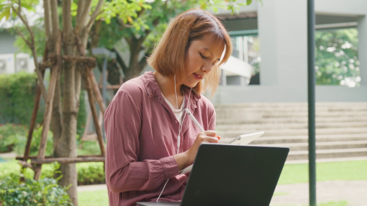 年轻的亚洲女孩戴着耳机，用笔记本电脑上网学习，坐在公园里在平板电脑上写字。视频素材