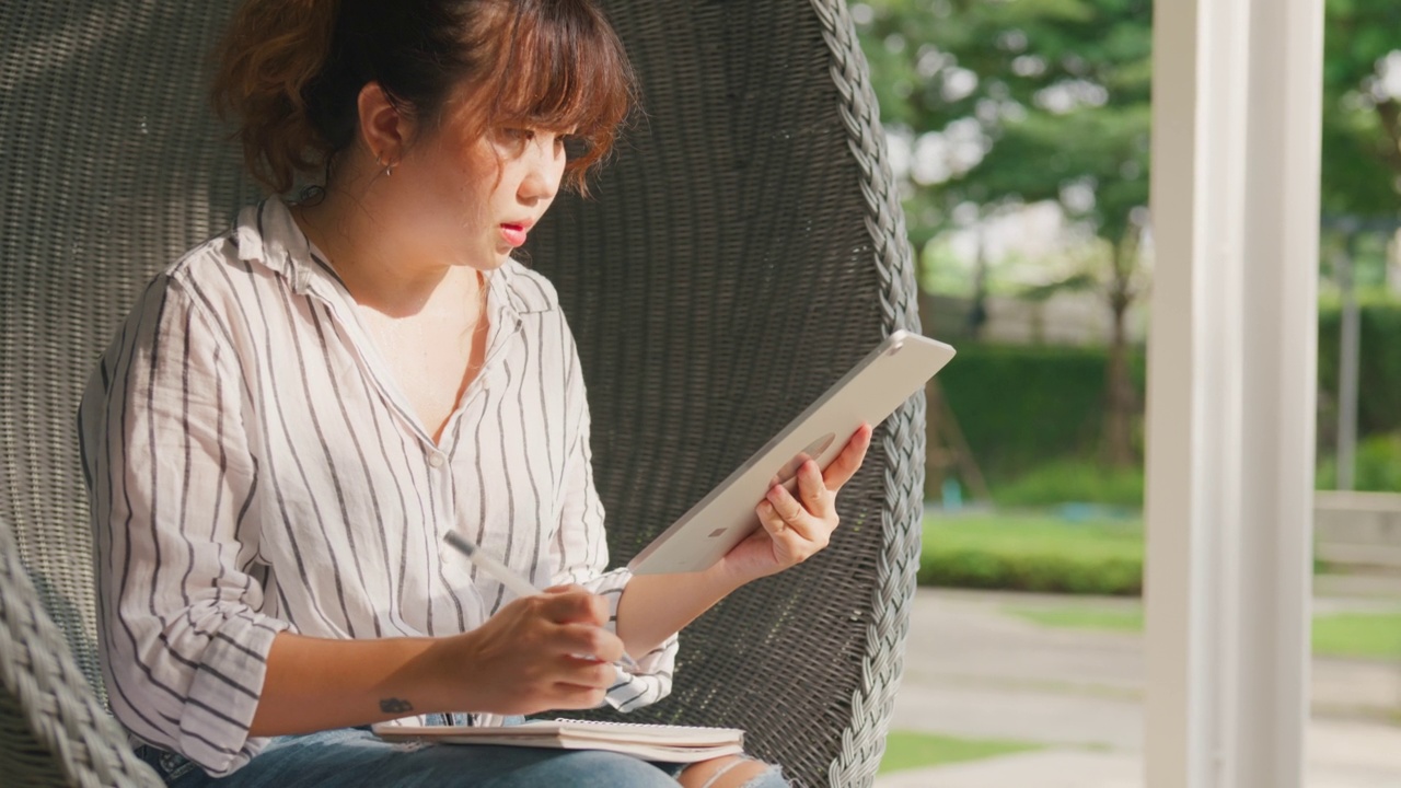 年轻的亚洲女孩坐在公园里用平板电脑在线学习。视频素材