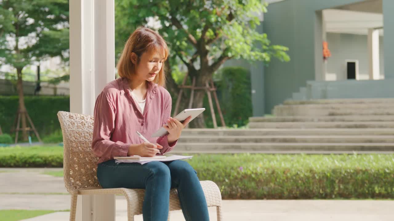 年轻的亚洲女孩坐在公园里用平板电脑在线学习。视频素材