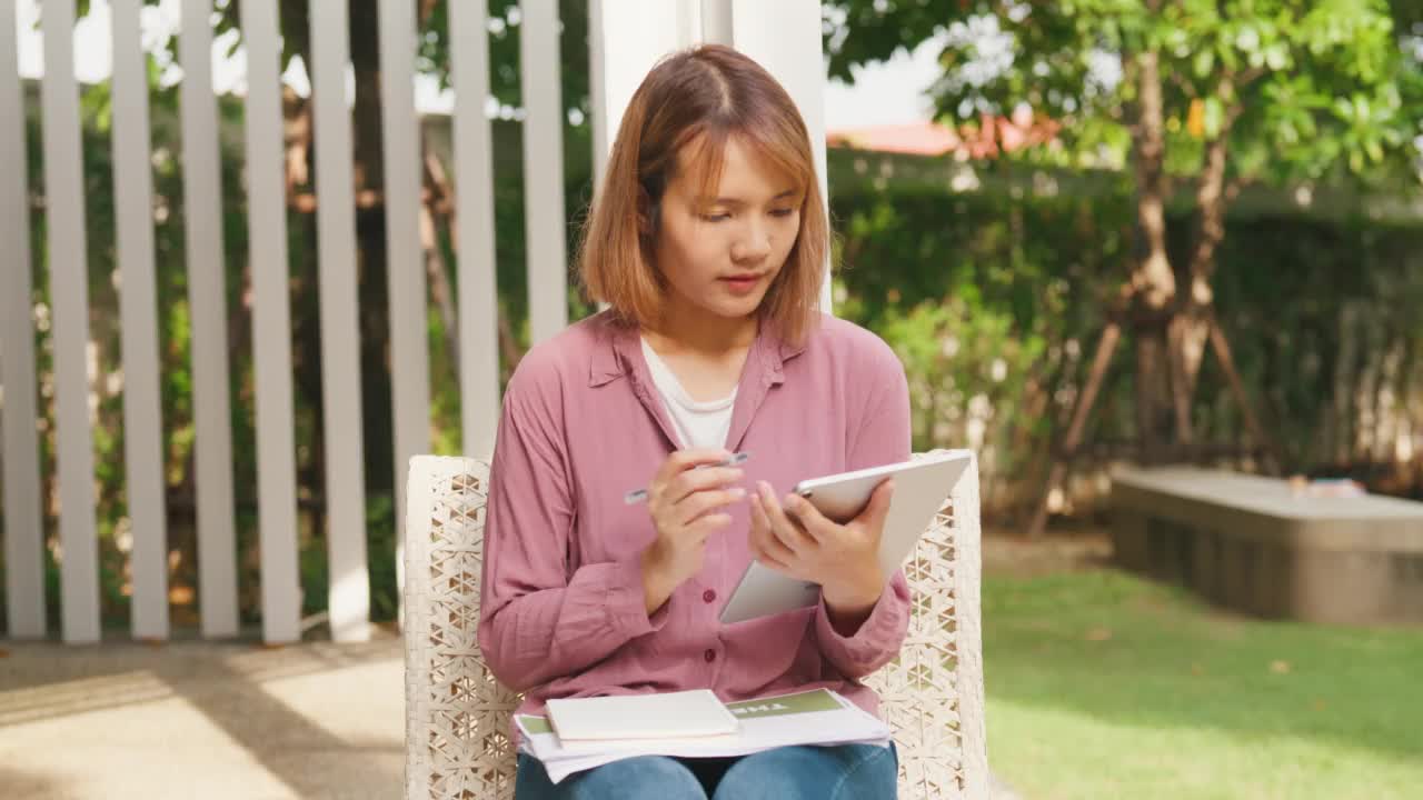 年轻的亚洲女孩坐在公园里用平板电脑在线学习。视频素材