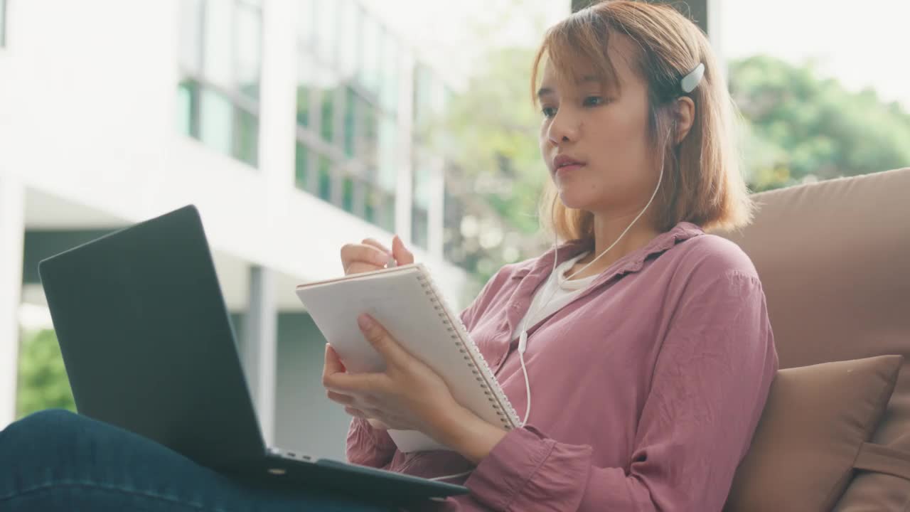 年轻的亚洲女孩戴着耳机用笔记本电脑上网学习，躺在游泳池附近的椅子上写笔记本。视频素材