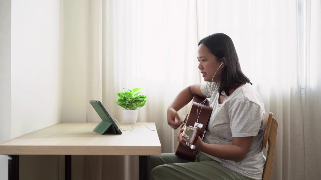 亚洲妇女练习玩木吉他在线使用平板电脑坐在家里靠近窗户的时候，在白天。以技能提升为理念的周末活动视频素材