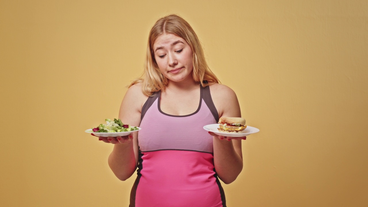 金发女人在健康食品和垃圾食品之间做选择。身穿粉色运动服的金发超重女性正在黄色背景下节食，在蔬菜沙拉和三明治之间做出选择视频素材