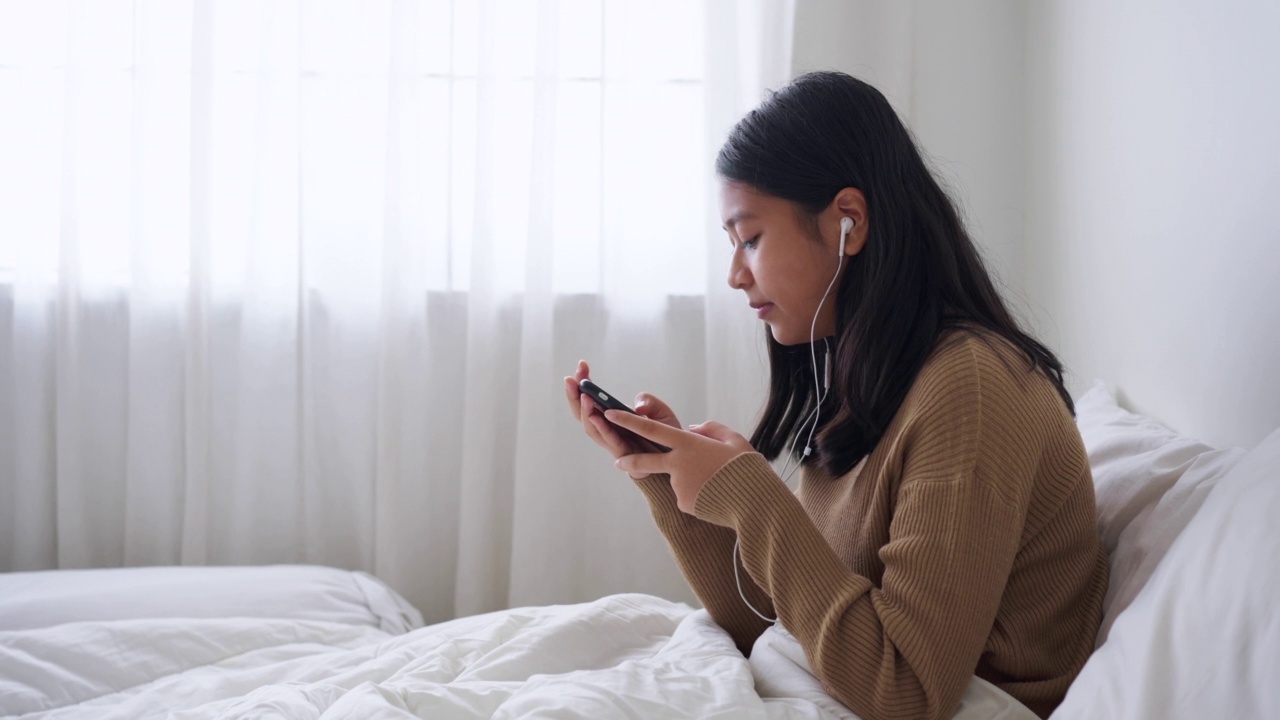 坐在床上的亚洲女孩喜欢在白天用智能手机在线听音乐视频素材