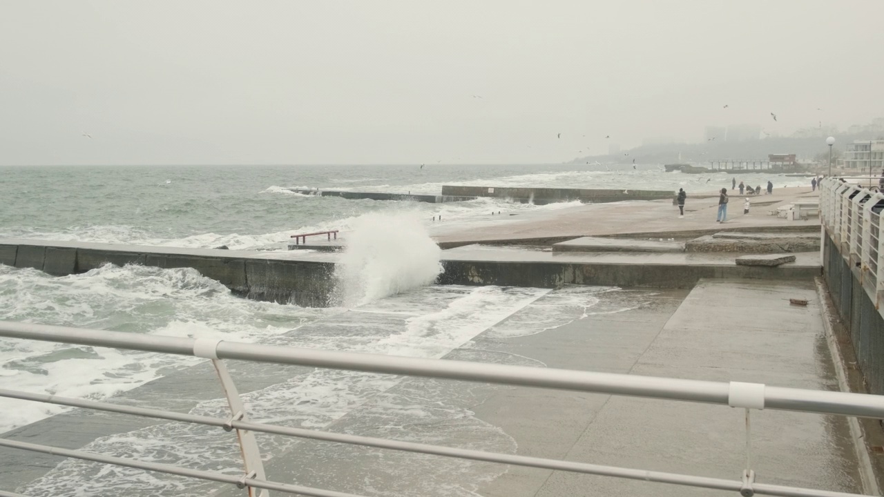 冬天的大海狂风大作，汹涌的海浪拍打着城市堤岸上的防波堤，天空雾蒙蒙的，人们穿着冬装在海滩上散步视频素材