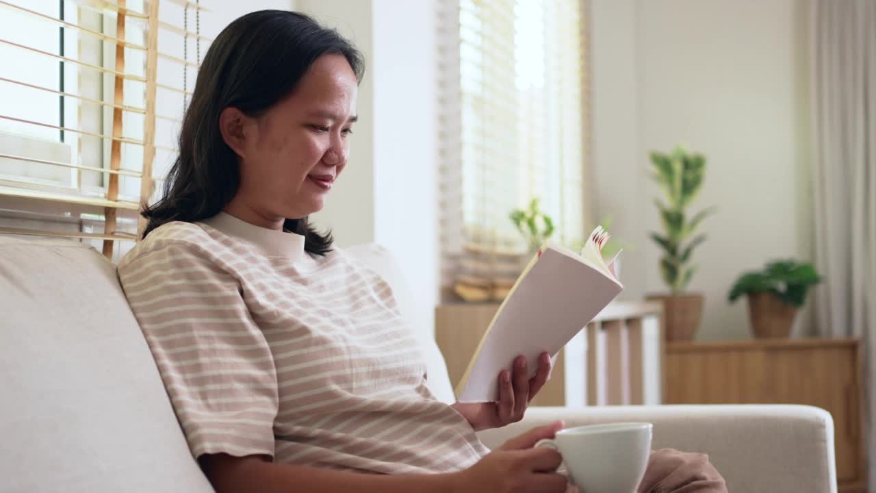 快乐的女人周末坐在沙发上看书，喝咖啡或茶在家里的客厅视频素材