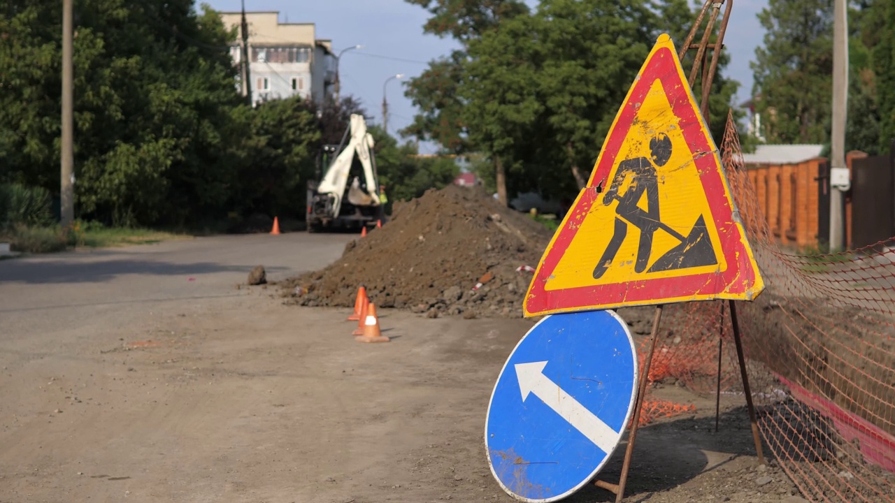 在建封闭道路上有箭头标志的警示标志，妨碍城市交通。背景上拖拉机推土机用铲斗收集泥土，倒在一大堆地上。视频素材