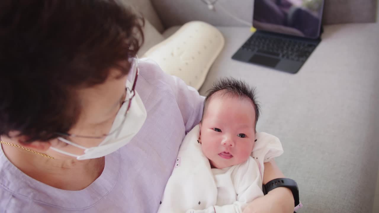 快乐的亚洲奶奶带着防护口罩说话看着孙女刚出生的婴儿婴儿在奶奶舒适的怀里在家里，退休的爷爷照顾孙子，童年视频素材