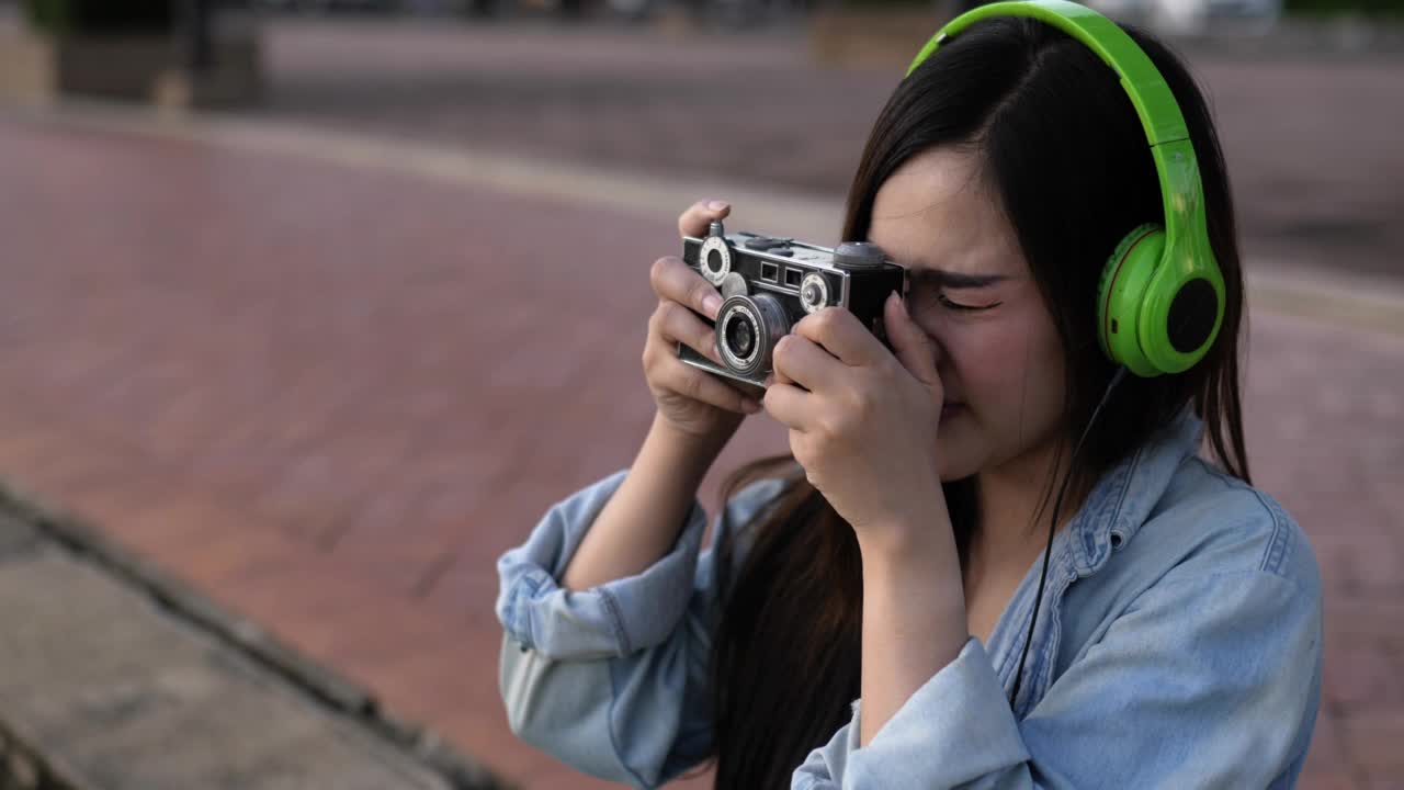 一名年轻女子戴着耳机在公园里听音乐，用相机拍照视频素材