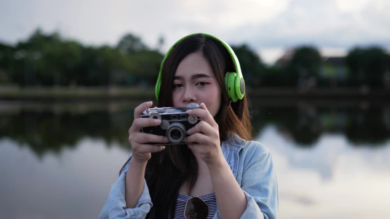 一名年轻女子戴着耳机在公园里听音乐，用相机拍照视频素材