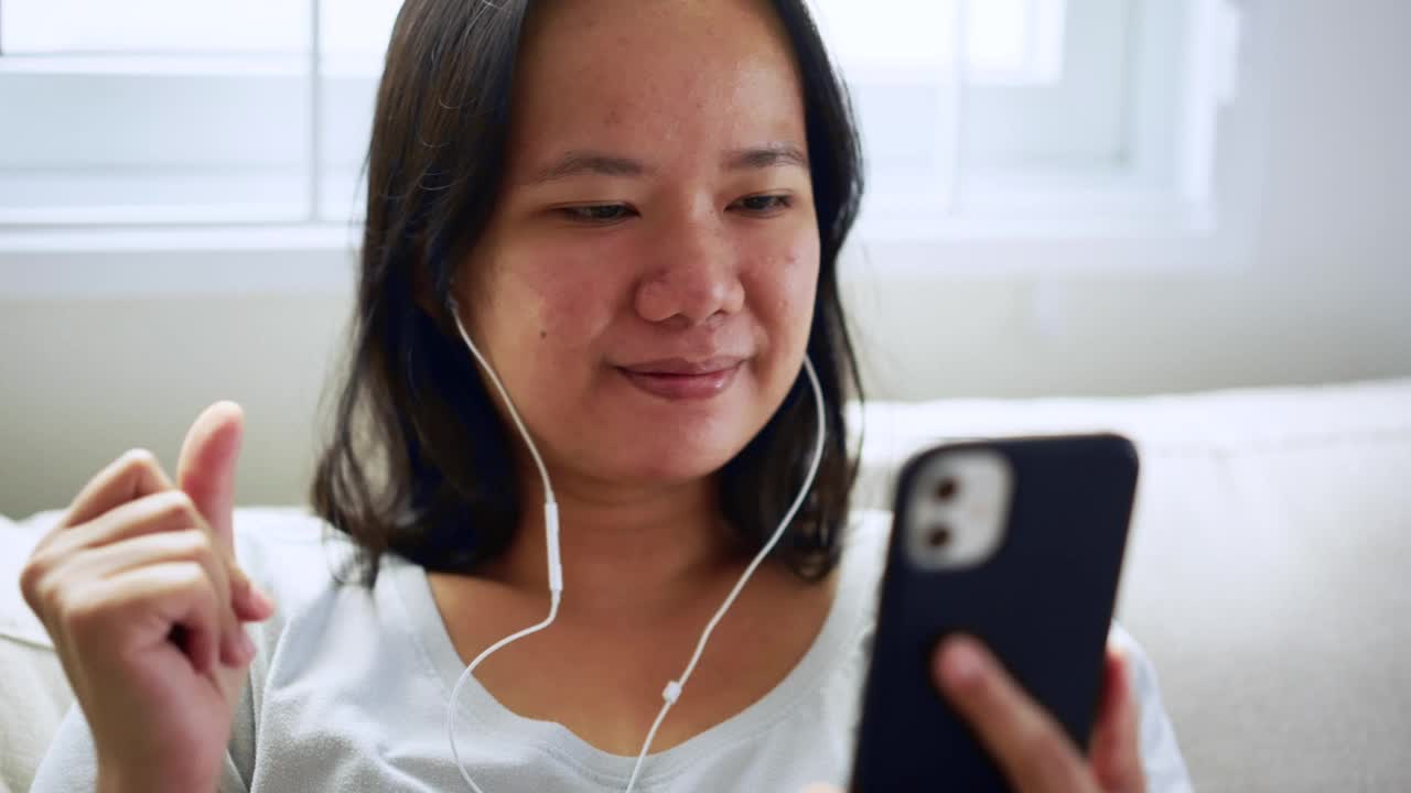 快乐的亚洲妇女坐在家里的沙发上，喜欢用手机听音乐。周末早上用智能手机放松视频素材