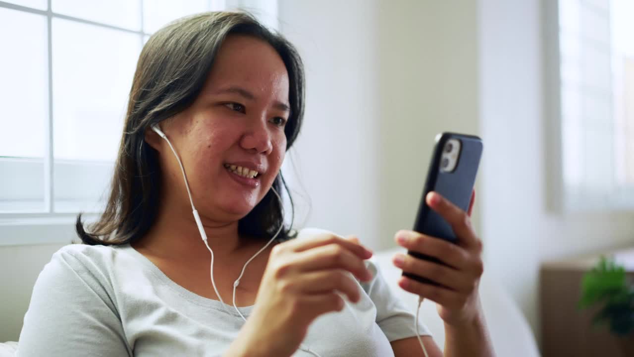 周末，戴着耳机的快乐亚洲女性喜欢坐在客厅沙发上用智能手机听音乐视频素材