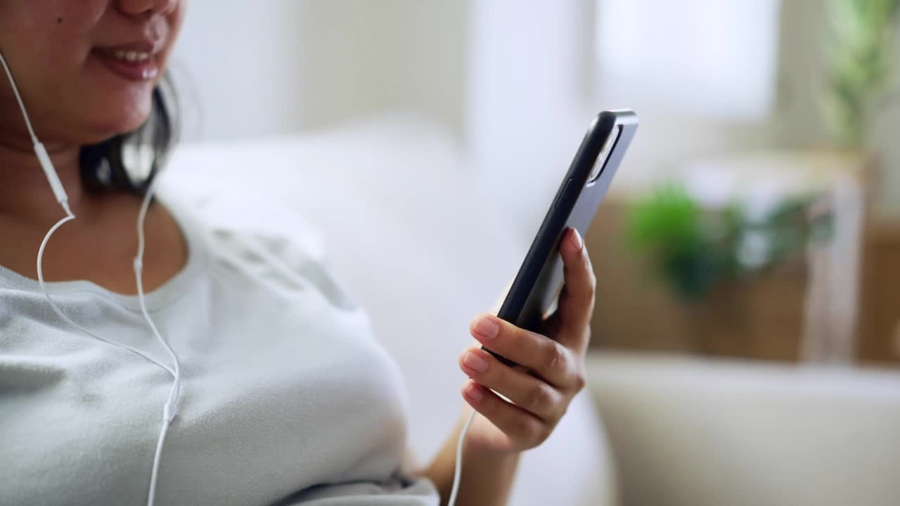 快乐的亚洲妇女坐在家里的沙发上，喜欢用手机听音乐。周末早上用智能手机放松视频素材