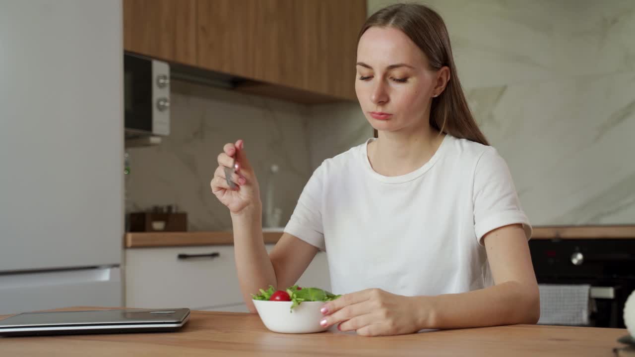 年轻的不满足的女人坐在家里的餐桌边吃沙拉视频素材