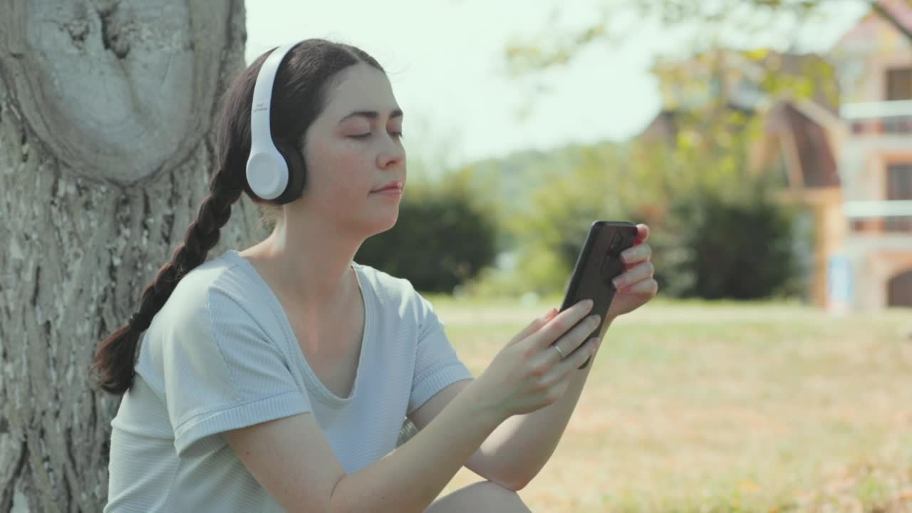 一个年轻漂亮的白人女子坐在公园里，戴着耳机，用着智能手机。用于教育和语言学习的移动应用程序视频素材