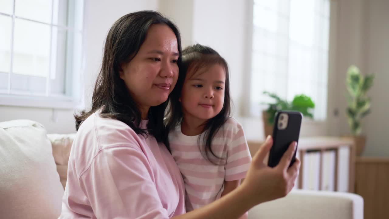 幸福的妈妈和女儿在家里的客厅里用智能手机微笑着进行在线视频通话。周末阖家欢乐视频素材