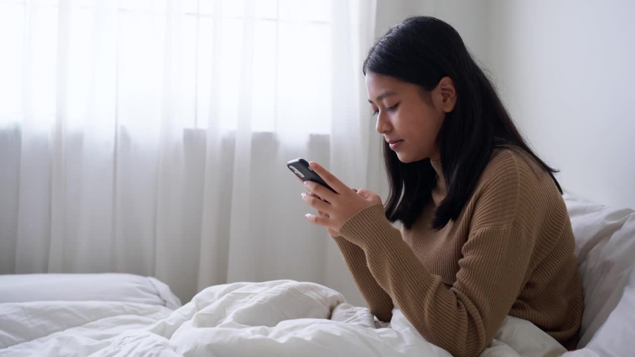 一个坐在床上的女孩喜欢在白天用智能手机发短信或上网聊天视频素材