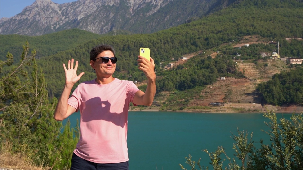 一名男子用手机自拍山脉湖泊背景。旅行者男在蓝湖户外旅行冒险假期用手机视频聊天。视频素材