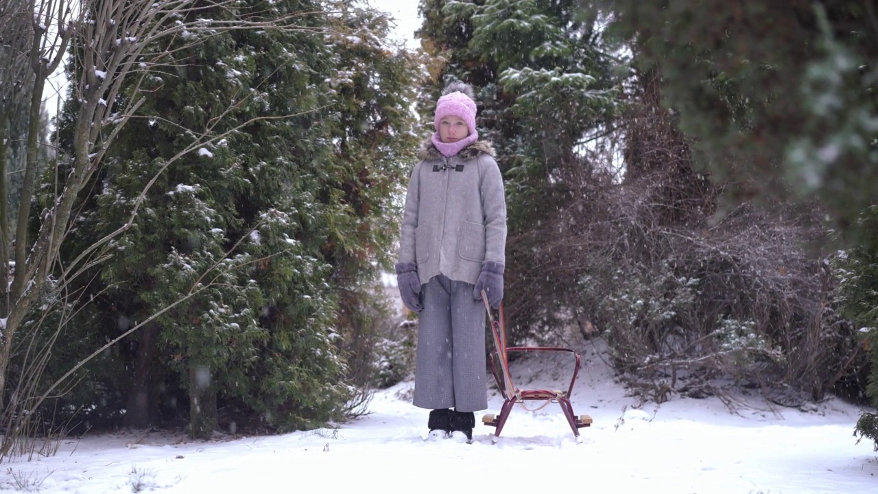 广角拍摄的肖像孤独的少女站在雪橇与雪花飘落在户外慢动作。悲伤的白人少年独自站在户外看着镜头。视频素材