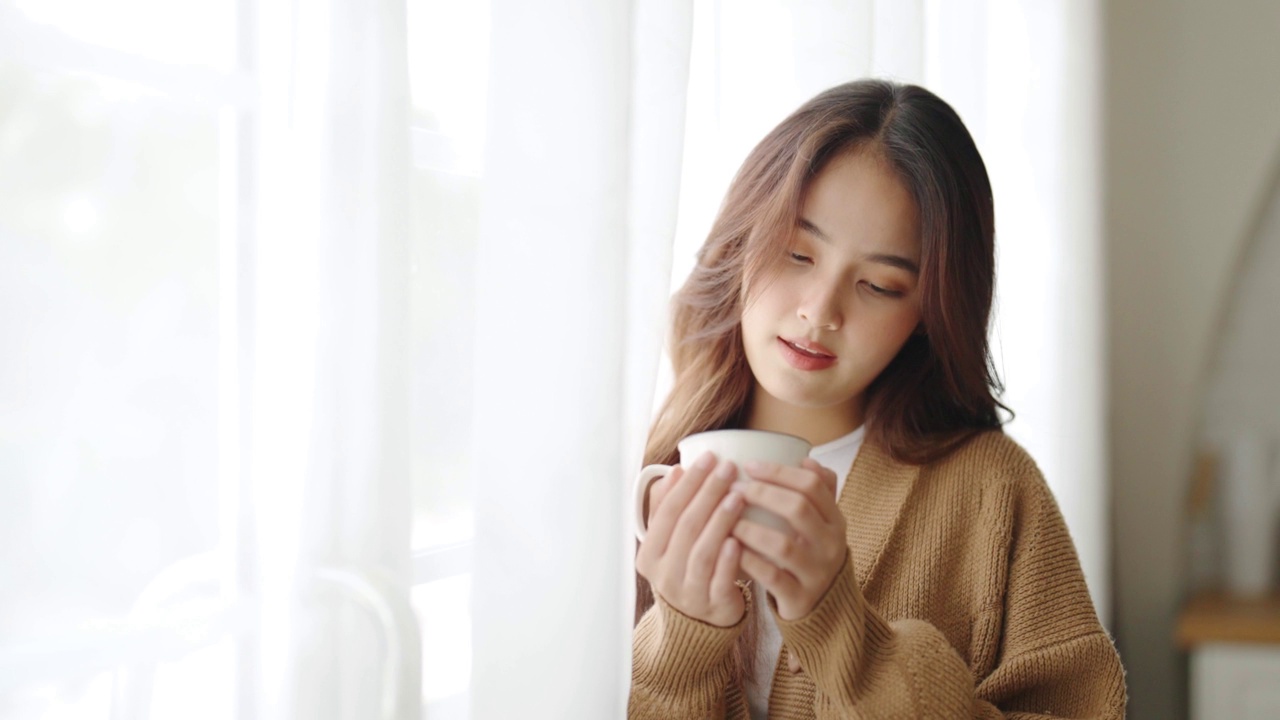 年轻的亚洲妇女站在窗户旁边，在家里的客厅拿着杯子。亚洲女性早上醒来后喝牛奶或茶视频素材