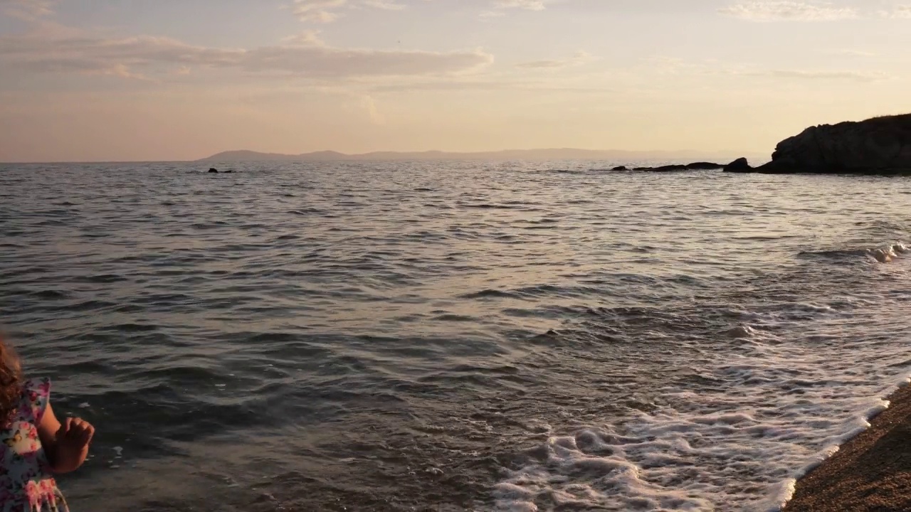 夕阳下的海边，母亲和蹒跚学步的女儿。母亲和小女孩走在被海浪拍打的海岸线上视频素材