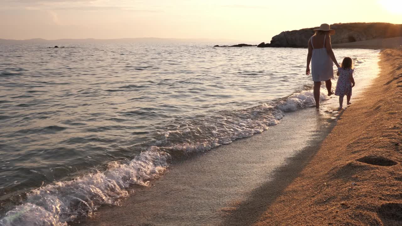 夕阳下的海边，母亲和蹒跚学步的女儿。母亲和小女孩走在被海浪拍打的海岸线上视频素材