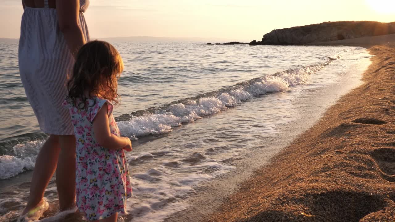 夕阳下的海边，母亲和蹒跚学步的女儿。母亲和小女孩走在被海浪拍打的海岸线上视频素材