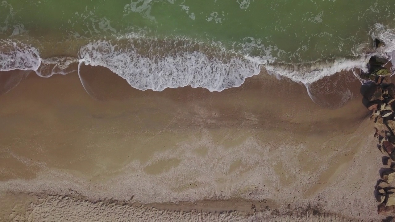 从上到下鸟瞰宁静的空海滩，泡沫的海浪冲刷海岸上的沙子。海浪冲击砂质涂层线上的石质防波堤。从上面看，无人机向左移动。视频素材