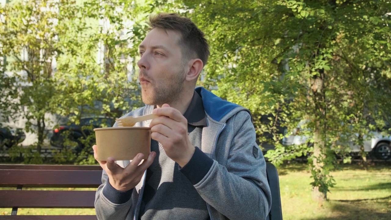 年轻的白人男子在公园里吃一次性纸包装或容器的沙拉。午餐盒里的健康食品视频素材